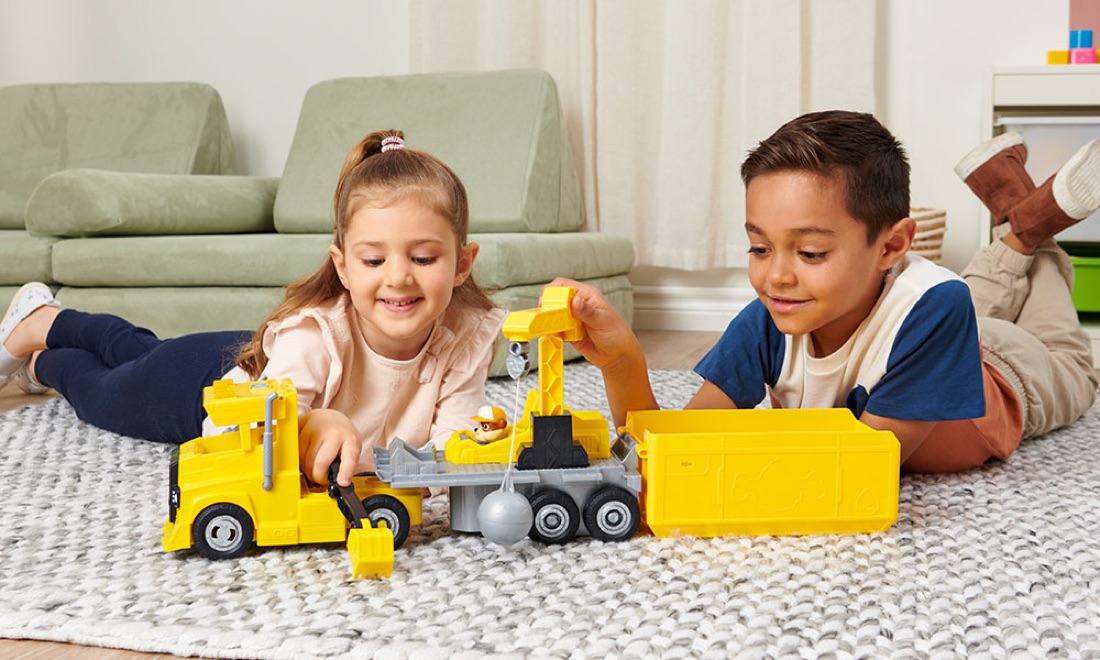 A girl and boy playing with a yellow toy truck on the living room floor