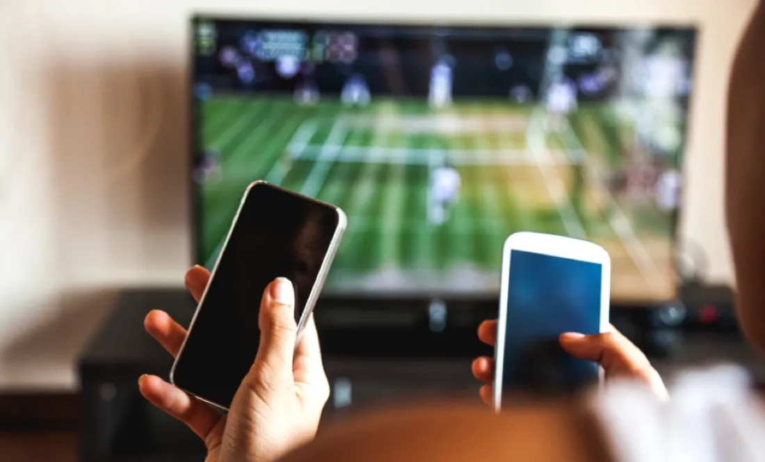 Two phones in hand in front of a TV showing live sports